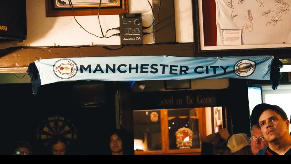 A Manchester City scarf is displayed in a pub, accompanied by a dartboard and autograph frames. The atmosphere suggests a sports-themed environment.