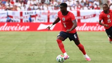 Soccer players in red jerseys playing on a field. Number 6 is dribbling the ball, with an English national emblem visible.