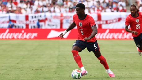 Soccer players in red jerseys playing on a field. Number 6 is dribbling the ball, with an English national emblem visible.