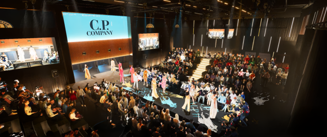 A fashion show event with models on a runway in front of an audience. The screen displays a logo 'C.P. Company' and multiple screens show different angles of the runway.