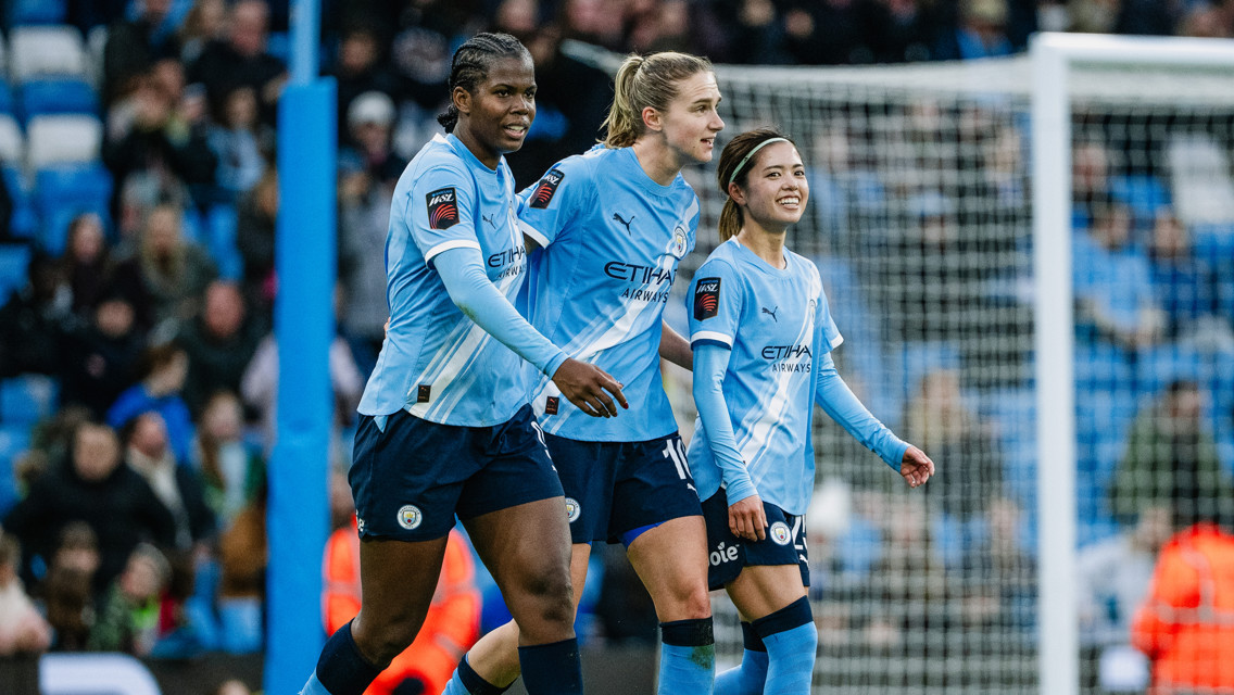 Three Manchester City women's football players walking on the pitch in their light blue kits during a match, with a blurred crowd in the background.
