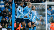 Three Manchester City women's football players walking on the pitch in their light blue kits during a match, with a blurred crowd in the background.