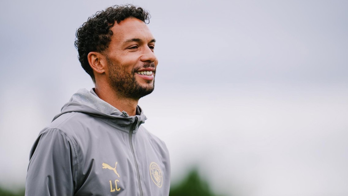 A smiling man wearing a grey jacket with initials 'LC' and the Manchester City logo, standing outdoors under a cloudy sky.