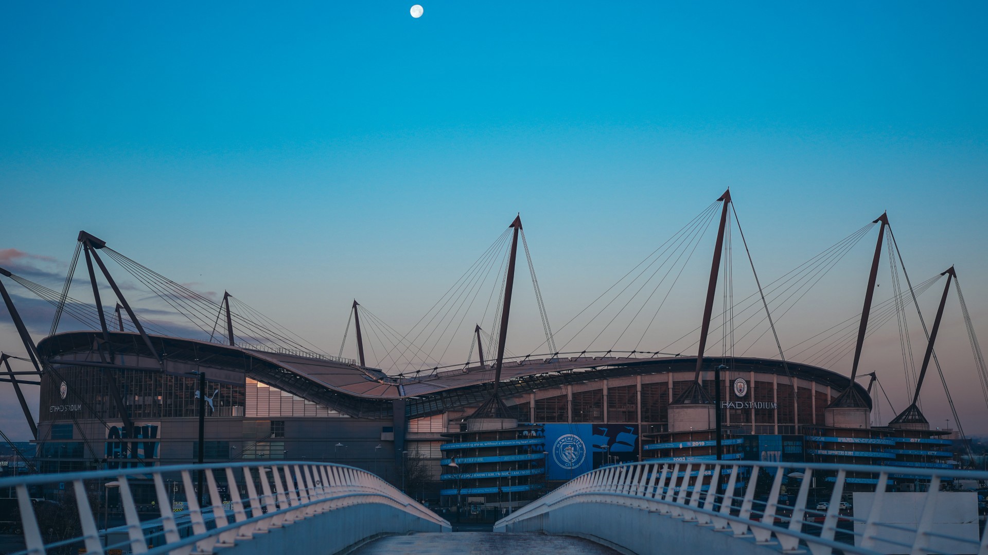 View of Etihad Stadium with a clear sky and the moon visible above. The modern architecture includes prominent lighting towers and a bridge leading towards the stadium.