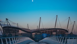 View of Etihad Stadium with a clear sky and the moon visible above. The modern architecture includes prominent lighting towers and a bridge leading towards the stadium.