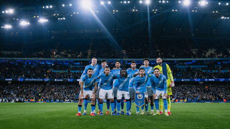 Group photo of Manchester City players in blue jerseys before a UEFA Champions League match at a stadium under bright lights.