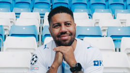 A person wearing a Manchester City jersey sitting in empty stadium seats.