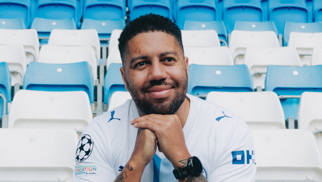 A person wearing a Manchester City jersey sitting in empty stadium seats.