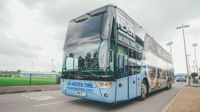 A large double-decker bus with Manchester City branding parked near a sports facility. The bus is predominantly blue with sponsors' logos, including Nexen Tire, and features the slogan 'We are City.'