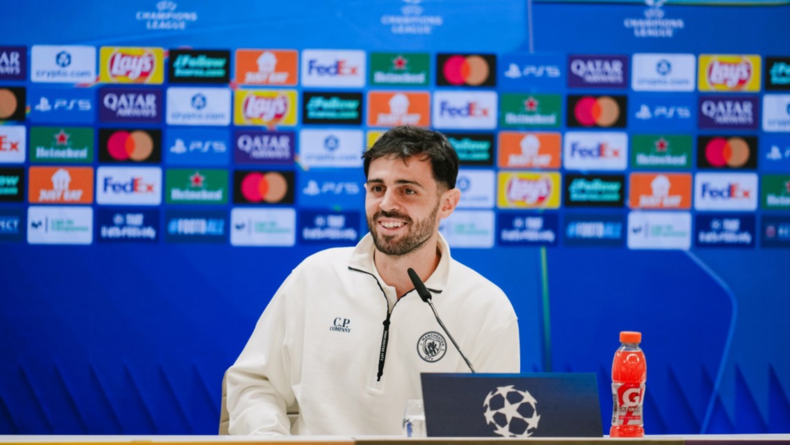 Blurred man seated at press conference with Champions League logos in background, wearing C.P. Company sweatshirt, microphone and Gatorade bottle visible.