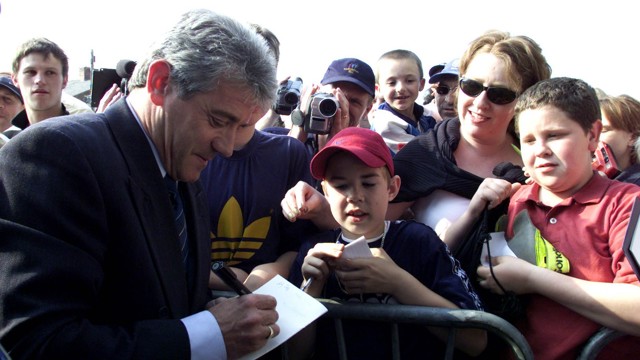 A man in a suit signing autographs for a group of fans behind a barrier. The fans include children and adults, some taking photos and videos.