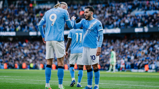 Manchester City players celebrating on the field in front of a crowd. Player wearing number 9 is interacting with player in number 10. Another player wearing number 11 is visible in the background.