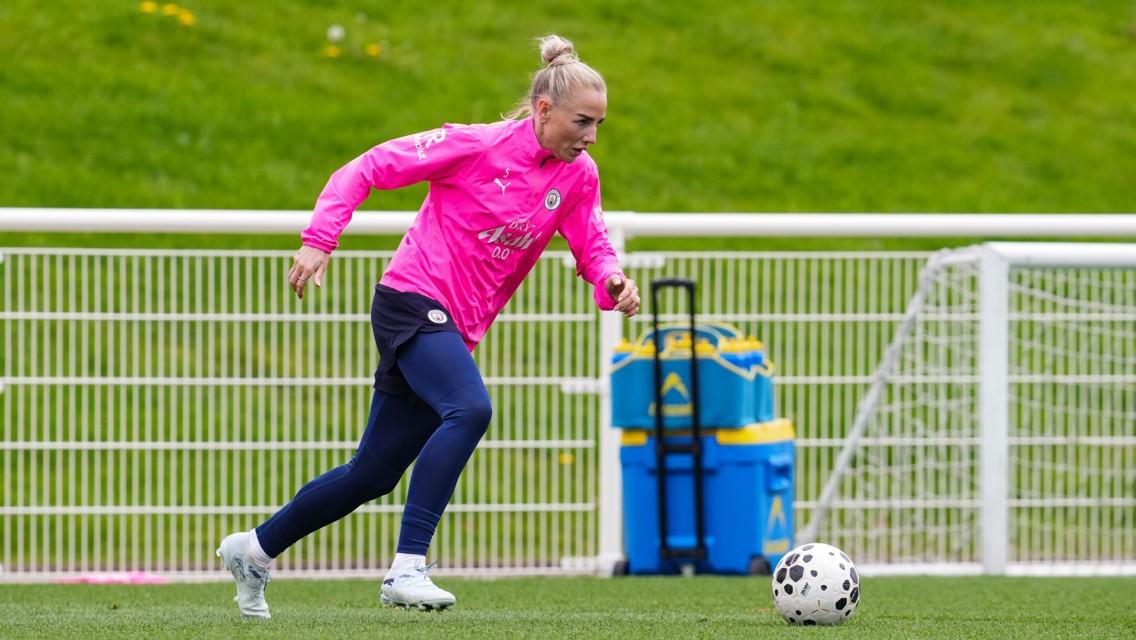A soccer player in a pink Manchester City training outfit dribbles a ball on the field with a goal net visible in the background.