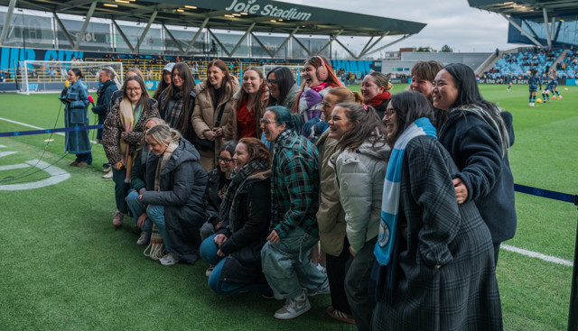 A group of people pose for a photo on the field of Joie Stadium, with part of a football pitch visible in the background.