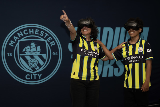 Two individuals wearing Manchester City jerseys pointing, with the Manchester City logo and badge in the background.