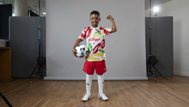 A young child stands in front of a gray backdrop wearing a colorful Kellogg's sports shirt, red shorts, white knee-high socks, and cleats, holding a soccer ball and flexing an arm. The face is blurred for privacy.
