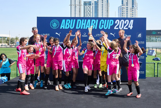 Youth soccer team wearing black and pink uniforms celebrates winning the Abu Dhabi Cup 2024, posing with medals and trophy at an award ceremony.