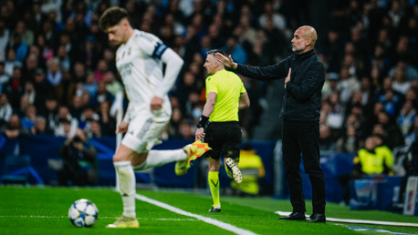 A soccer coach giving instructions from the sideline while a player runs with the ball and a referee is nearby. The setting is a stadium during a match.