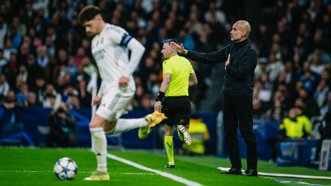 A soccer coach giving instructions from the sideline while a player runs with the ball and a referee is nearby. The setting is a stadium during a match.