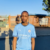 A person wearing a Manchester City jersey with the Etihad Airways logo standing outside in front of a gated area under a clear blue sky.