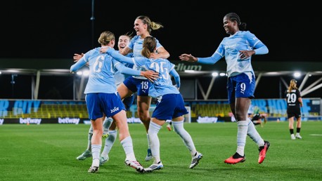 A group of Manchester City women's football players celebrate a goal on the pitch, wearing light blue and dark blue kits with sponsorship logos visible. The stadium lights illuminate the scene during a nighttime match.