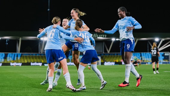 A group of Manchester City women's football players celebrate a goal on the pitch, wearing light blue and dark blue kits with sponsorship logos visible. The stadium lights illuminate the scene during a nighttime match.