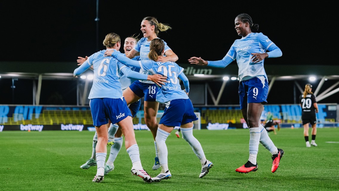 A group of Manchester City women's football players celebrate a goal on the pitch, wearing light blue and dark blue kits with sponsorship logos visible. The stadium lights illuminate the scene during a nighttime match.