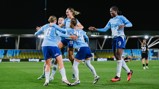 A group of Manchester City women's football players celebrate a goal on the pitch, wearing light blue and dark blue kits with sponsorship logos visible. The stadium lights illuminate the scene during a nighttime match.