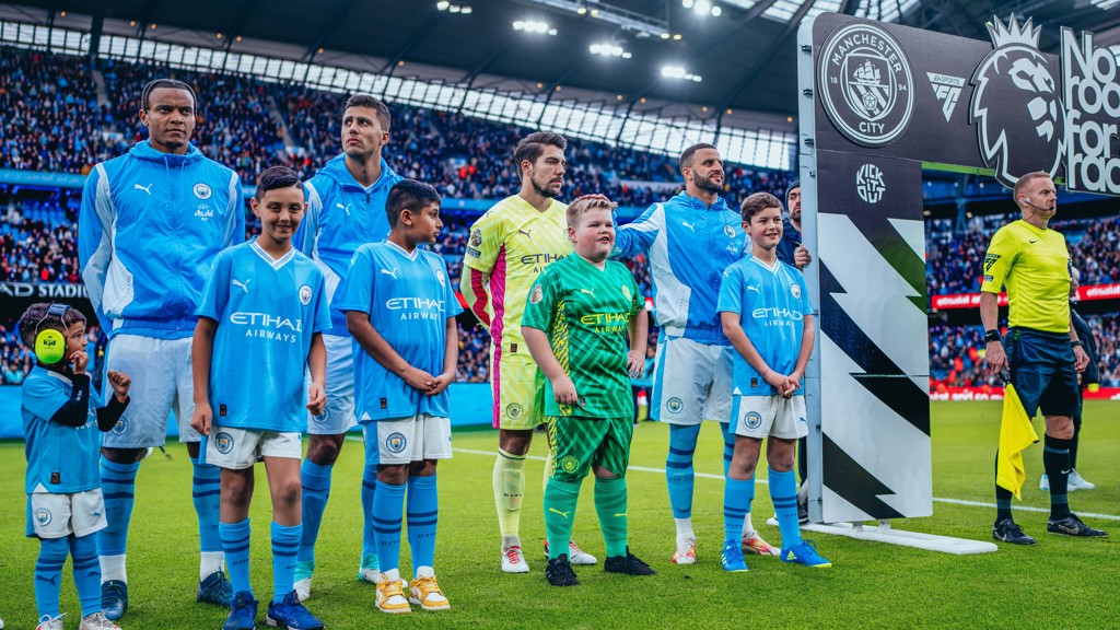 Manchester City players and young mascots line up on the pitch at the Etihad Stadium before a match. The Manchester City logo and the Premier League logo are displayed prominently. One mascot is seen in a full kit and headset.
