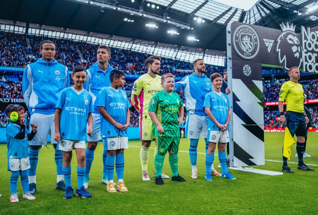 Manchester City players and young mascots line up on the pitch at the Etihad Stadium before a match. The Manchester City logo and the Premier League logo are displayed prominently. One mascot is seen in a full kit and headset.