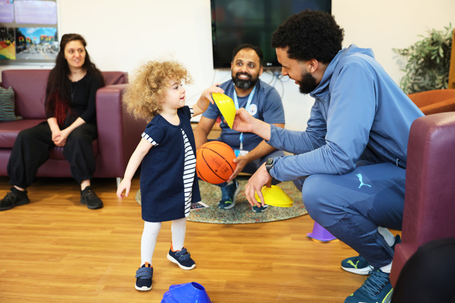 A young child interacting with a coach in a sports session, surrounded by adults. The child is handing a yellow cone to the coach, who is holding more cones. A basketball is visible in the background.