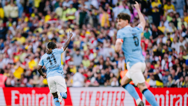 Two Manchester City players celebrate a goal in front of a cheering crowd. One player is wearing the number 11 jersey, pointing upwards.