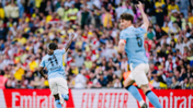 Two Manchester City players celebrate a goal in front of a cheering crowd. One player is wearing the number 11 jersey, pointing upwards.