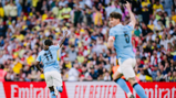 Two Manchester City players celebrate a goal in front of a cheering crowd. One player is wearing the number 11 jersey, pointing upwards.