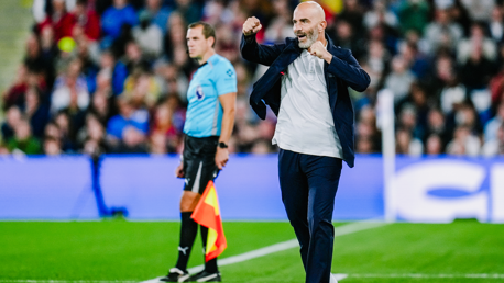 A soccer coach on the sideline gesturing during a match. An assistant referee in uniform is visible in the background.