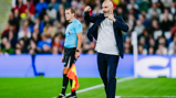 A soccer coach on the sideline gesturing during a match. An assistant referee in uniform is visible in the background.
