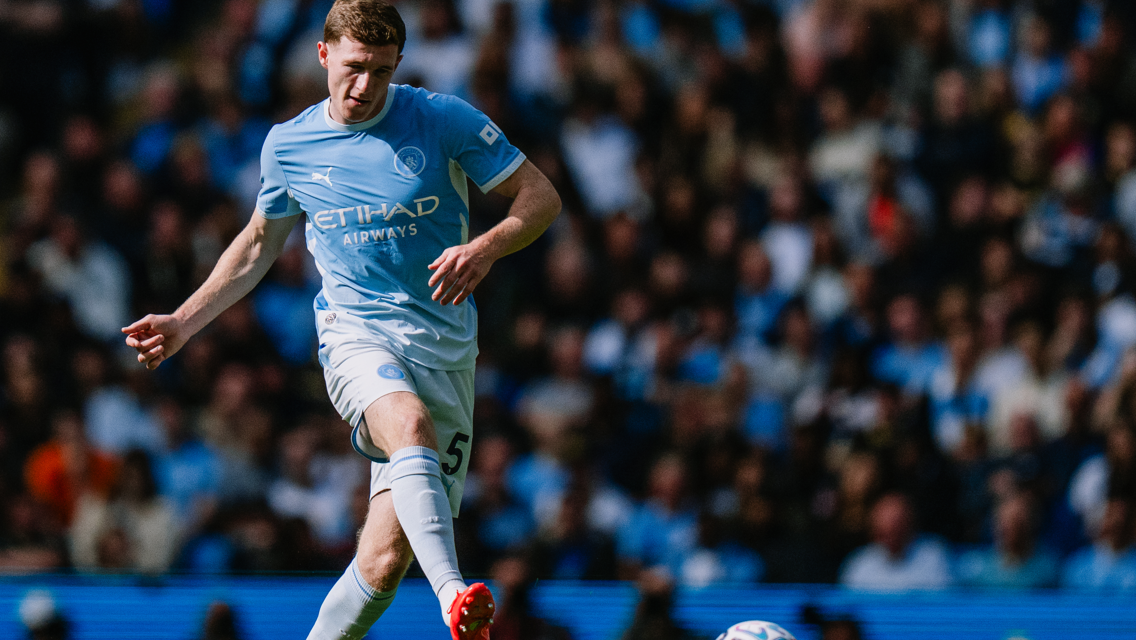 A Manchester City player wearing a light blue kit is in action on the field, with a blurred crowd in the background.