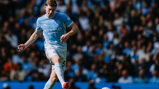 A Manchester City player wearing a light blue kit is in action on the field, with a blurred crowd in the background.