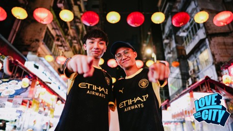 Two people in Manchester City jerseys point at the camera while standing under colorful lanterns in a lively street. 'Love This City' is visible in the corner.