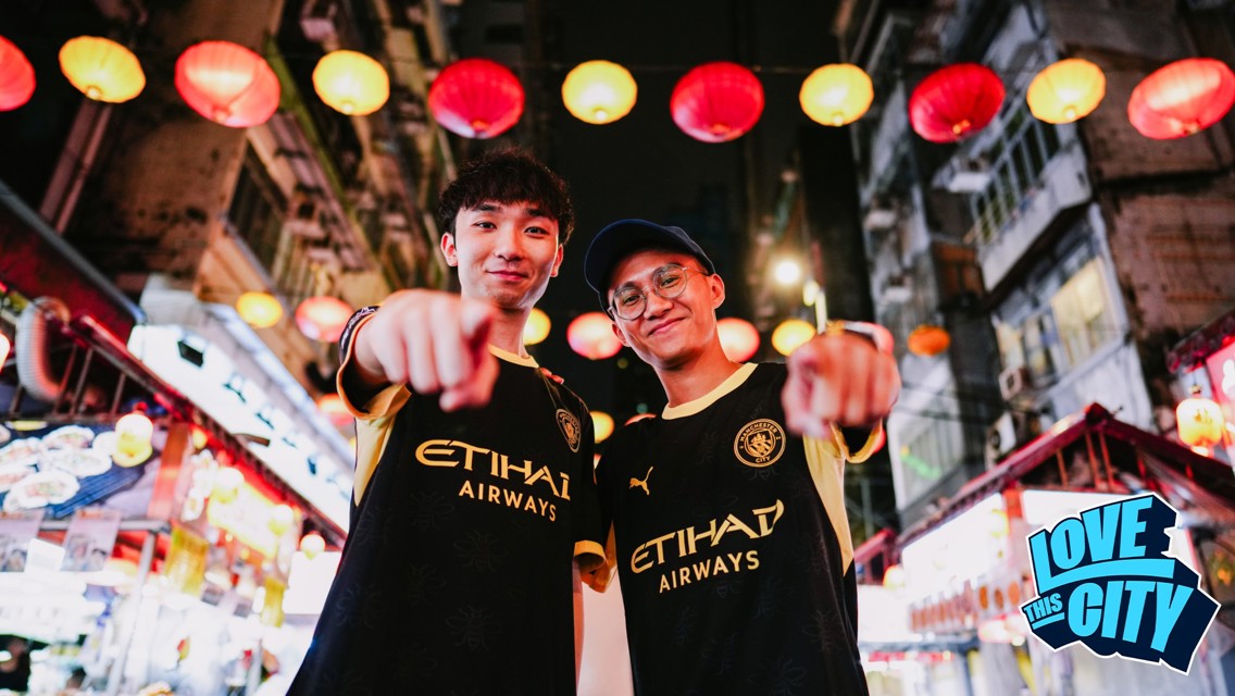 Two people in Manchester City jerseys point at the camera while standing under colorful lanterns in a lively street. 'Love This City' is visible in the corner.