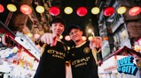 Two people in Manchester City jerseys point at the camera while standing under colorful lanterns in a lively street. 'Love This City' is visible in the corner.