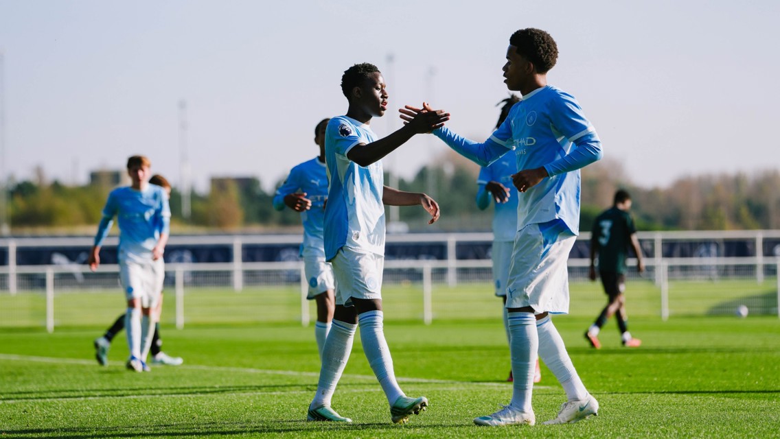 Two young football players in light blue kits celebrating on a football field by shaking hands, with other players in the background.