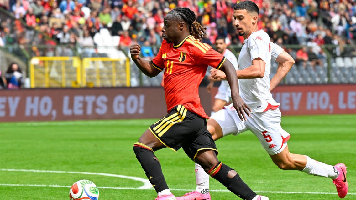 Soccer match between Belgium and Switzerland players, with Belgium player dribbling the ball and Swiss defender in pursuit. Crowd is visible in the background.