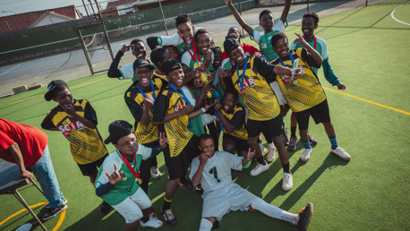A group of young soccer players wearing medals and uniforms celebrate with a trophy on a sports field.