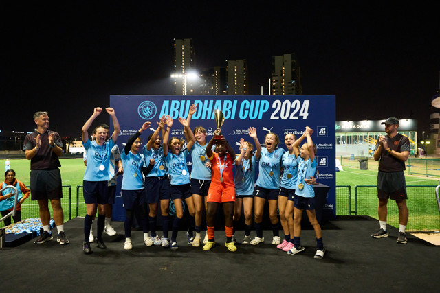 A team of young female football players celebrating with a trophy on the podium at the Abu Dhabi Cup 2024.