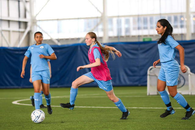 Three girls playing soccer indoors, one wearing a pink vest and the other two in blue uniforms. A ball is on the field and they are actively engaged in the game.