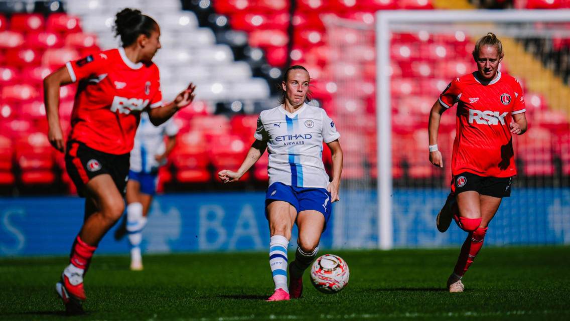 A women's football match featuring Manchester City wearing white and blue uniforms against Charlton Athletic in red, in a stadium with red and white seating.