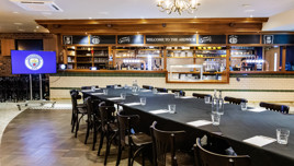 A conference room with a long table set up, chairs, and water glasses. The room is decorated with a Manchester City logo on a TV screen. The bar area is visible in the background.
