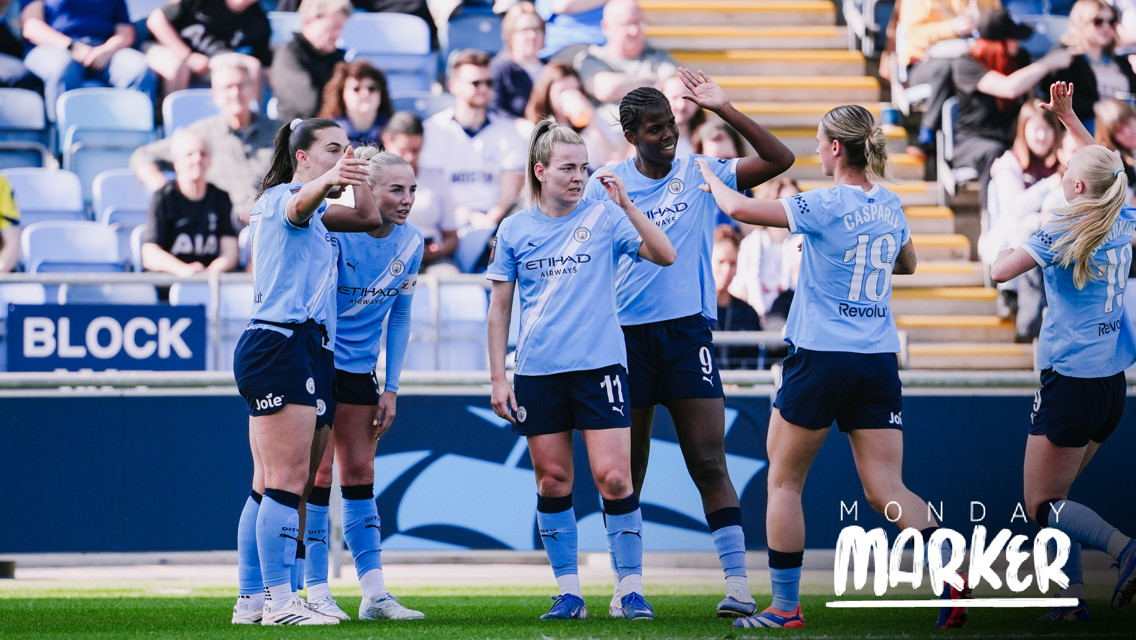 Group of Manchester City women's football team players celebrating on the field, wearing light blue kits in front of a crowd.