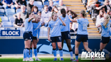 Group of Manchester City women's football team players celebrating on the field, wearing light blue kits in front of a crowd.